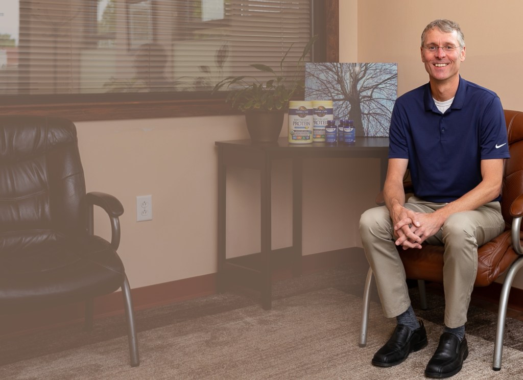 Dr. Bart Hodgins, a chiropractor in Asheville, NC, smiling in his wellness office next to nutrition supplements.