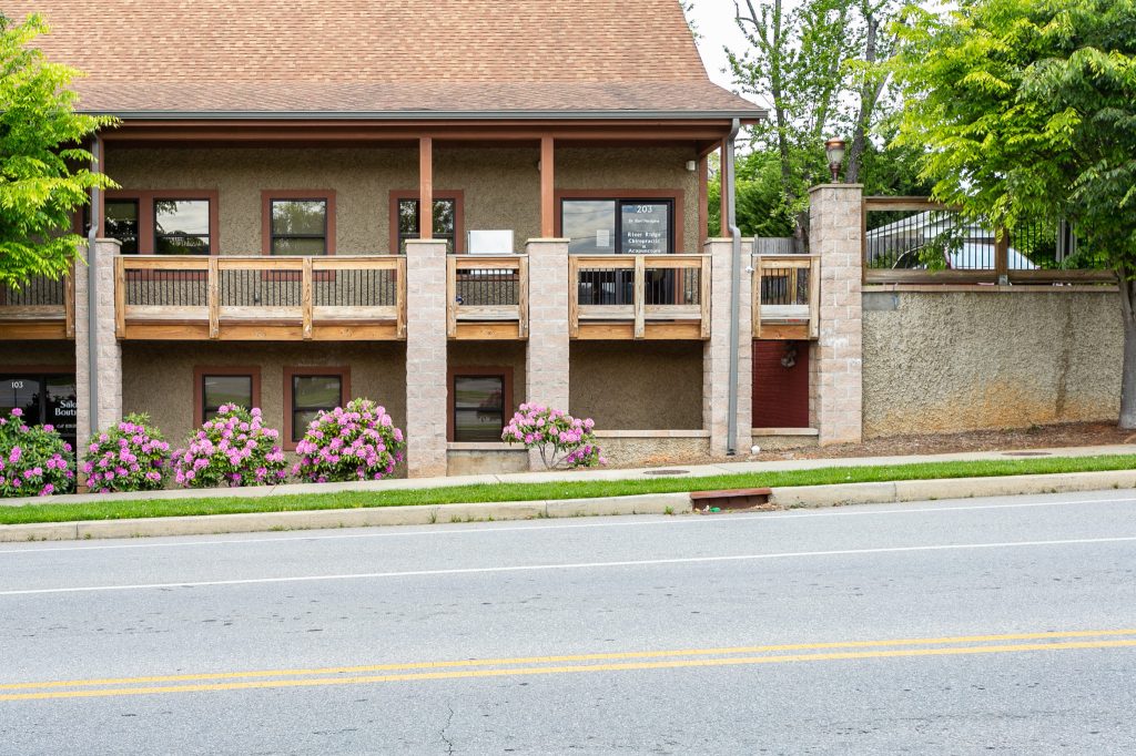 Exterior street-view of the River Ridge Chiropractic & Acupuncture office building in Asheville, featuring a second-floor wooden balcony and pink rhododendron bushes.
